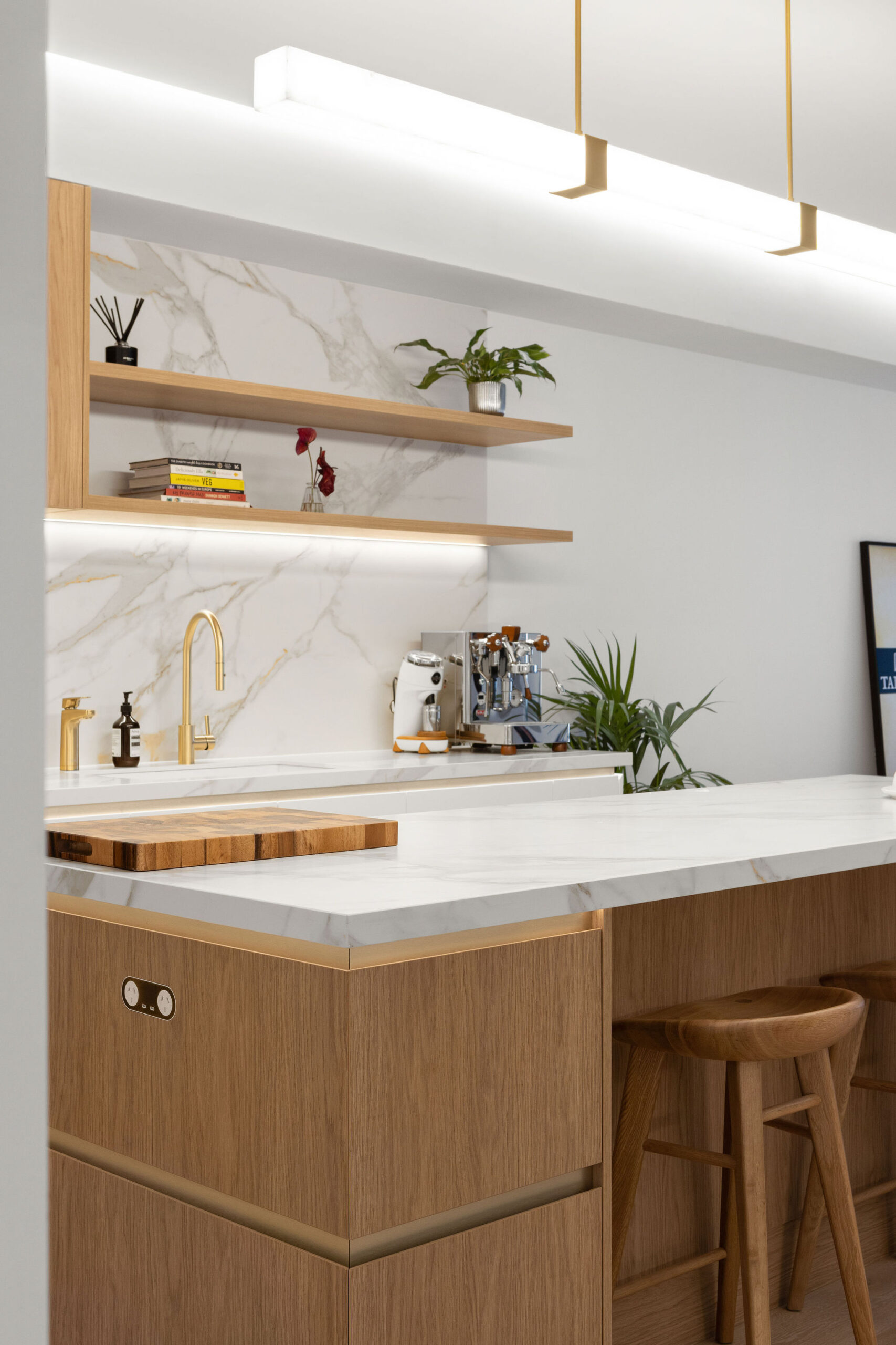 Perspective view of island bench in kitchen looking across to overhead open shelves. Woodgrain finishes with stone benchtops.